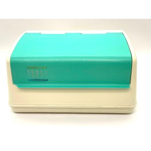 May include: A rectangular bread box with an off-white base and a hinged teal lid. The front of the teal lid features the word BREAD printed in a stylized, outlined font, with a decorative horizontal line graphic positioned directly below the text.