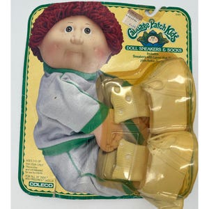 May include: A Cabbage Patch Kids doll accessory set in original packaging, featuring yellow sneakers and socks. The cardboard backing displays the brand logo, the text Doll Sneakers & Socks, and includes a graphic of a doll with red yarn hair.