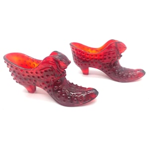 May include: Two translucent red glass shoe figurines featuring a hobnail texture across the exterior surface. Each decorative shoe has a pointed toe, a small raised heel, and a smooth opening at the top, showcasing a deep ruby red color throughout.