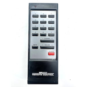 May include: A black rectangular remote control with a silver faceplate featuring a grid of twelve lettered buttons labeled A through L. It includes black rocker buttons for channel and volume control, a mute button, and a red power button labeled POWER.