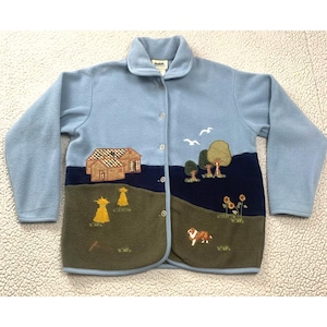 May include: A light blue fleece jacket featuring a button-down front and a landscape scene. The design includes embroidered houses, trees, sunflowers, haystacks, and a dog, with two white birds flying in the sky against a dark blue background.
