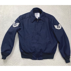 May include: A dark navy blue zip-up jacket featuring a pointed collar, ribbed cuffs, and a ribbed hem. Each sleeve displays a white and navy blue embroidered chevron patch with a central star. A white label inside the collar reads COYOTE.