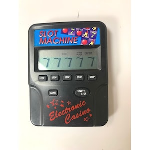 May include: A black handheld electronic slot machine game featuring a digital LCD screen displaying five sevens and the text 100 CREDIT. The device includes five stop buttons, a sound button, a start/stop button, and the text SLOT MACHINE and Electronic Casino.
