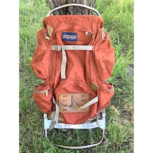 May include: A rust-orange JanSport external frame backpack featuring a metal frame, multiple zippered side pockets, a top flap, and a tan leather patch on the lower front with the letters JS embossed. The pack includes light tan adjustable straps.