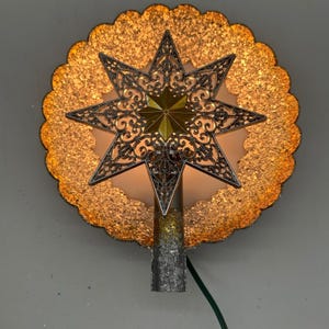Könnte beinhalten: Ein beleuchteter Baumspitzen-Aufsatz mit einer kreisförmigen, goldfarben glitzernden Scheibe mit gewelltem Rand und einem zentralen, filigranen, dunkelmetallenen achtstrahligen Stern. Ein kleinerer, massiver Goldstern liegt in der Mitte. Zylindrischer Sockel.