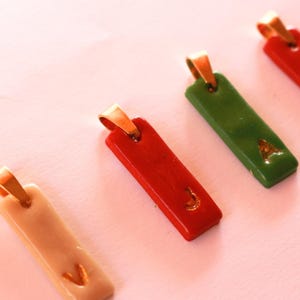 May include: A collection of rectangular pendants featuring a smooth, glossy finish in various colours including beige, red, and green. Each pendant is topped with a gold-toned metal bail and displays a single embossed letter in a gold-toned finish.