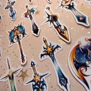 May include: A set of 10 colourful stickers featuring various sword designs with a celestial theme. The swords are decorated with stars, moons, and galaxies. The stickers are perfect for adding a touch of fantasy to your planner, laptop, or water bottle.