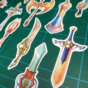 May include: A collection of colorful fantasy sword stickers with intricate details. The stickers feature various designs, including a golden axe, a blue and gold sword with a sun design, and a green and red sword with a crystal tip.