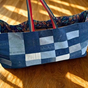 May include: A large denim tote bag with a patchwork design in various shades of blue. The bag features a floral patterned lining and red and blue striped handles. The bag is sitting on a wooden floor.