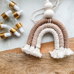 May include: A macrame rainbow wall hanging featuring three arched layers in shades of tan, cream, and white with fringed ends, topped with a wooden bead and a felt ball on a hanging cord, alongside four small glass dropper bottles with gold caps.