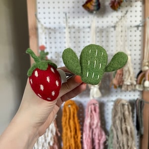 May include: A hand is holding two felt ornaments: a red strawberry with white seeds and a green cactus with white stitching.