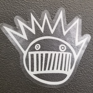 May include: White sticker with a cartoon face with a jagged crown and a mouth with teeth. The face has two eyes with pupils.