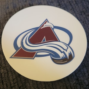 May include: A white round coaster with a blue and maroon logo of the Colorado Avalanche hockey team. The logo features a stylized "A" with a wave design.