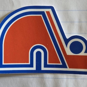 May include: A blue and white sticker with a red, white, and blue hockey logo. The logo is a stylized letter "A" with a circle on the right side.