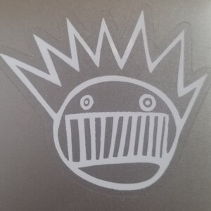 May include: A white sticker with a cartoon face with a mouth full of teeth and two eyes. The face has a crown of spikes on top of its head.