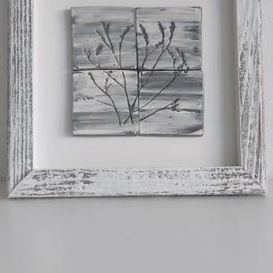 Può includere: Un'opera d'arte incorniciata con un design botanico. L'opera è composta da quattro piastrelle quadrate disposte all'interno di una cornice di legno bianco invecchiato. Le piastrelle presentano uno schema di colori grigio e bianco con un'illustrazione botanica nera.