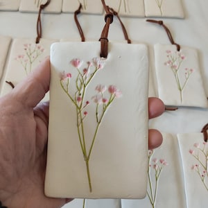May include: Rectangular white ceramic tags with pressed flowers and brown leather cords. One tag is held in a hand, displaying a sprig of pink baby's breath. Other tags feature lavender and baby's breath designs.