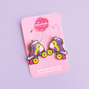 May include: A pair of gold hoop earrings with colourful roller skate charms. The skates are purple, pink, yellow, and green with white wheels. The earrings are on a pink card with the text "Studio Evannave" and "@evannaveillustration www.evannave.co.uk".