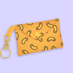 May include: A yellow key chain with a small pouch featuring a repeating pattern of macaroni. The pouch has a yellow button closure and a gold metal key ring.