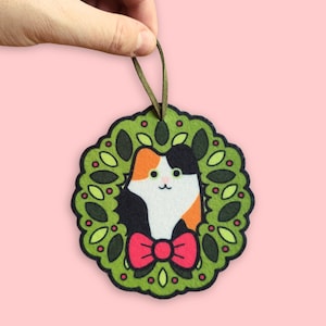 May include: A felt cat decoration featuring a green wreath design. The cat has black, orange, and white fur and a red bow tie. The ornament is held by a brown ribbon against a pink backdrop.