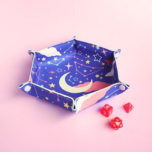 May include: A blue and pink dice tray with a celestial pattern featuring stars, a crescent moon, and constellations. The tray is made of fabric and has a white border. Three red dice are on a pink surface next to the tray.