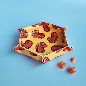 May include: A yellow dice tray with a white border and a red and orange dragon pattern. The tray is made of felt and has a hexagonal shape. There are four orange dice on the table next to the tray.