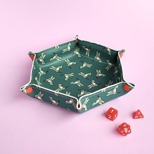 May include: A green felt dice tray with a hummingbird pattern and red button accents. Three red dice are on a pink surface next to the tray.