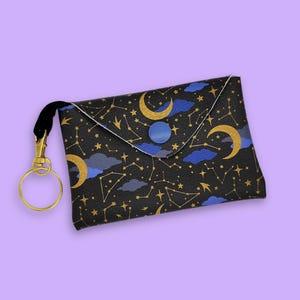 May include: Black clutch bag with a celestial design featuring gold moons, stars, and constellations, with blue accents. The bag has a gold-coloured clasp and a black wrist strap. A blue button secures the flap.