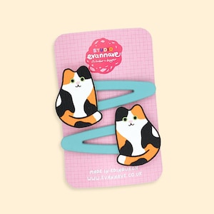 May include: Two cat-shaped hair clips with orange, black, and white markings, resting on light blue snap clips. The clips are on a pink card with the text "Studio Evannave" and "Made in Edinburgh".