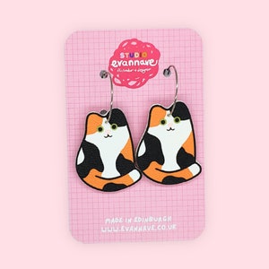 May include: A pair of hoop earrings featuring a calico cat design. The earrings are made of wood and have a black, orange, and white color scheme. The earrings are on a white card with the text "Studio Evannave" and "Handmade + Design" printed on it.