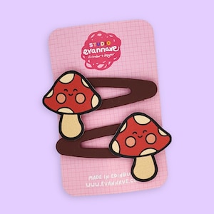 May include: Two red mushroom-shaped hair clips with white spots and smiling faces, attached to brown barrettes. The clips are on a pink card with text and a logo, set against a light purple background.