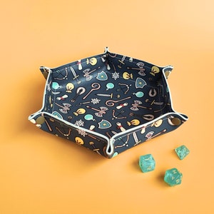 May include: A hexagonal dice rolling tray with a dark blue fabric interior patterned with fantasy game icons. The tray has a white trim and is secured with snap closures. Three green dice are scattered nearby.