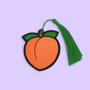 May include: A peach-shaped bookmark with a green tassel. The peach is orange with a black outline and green leaves.
