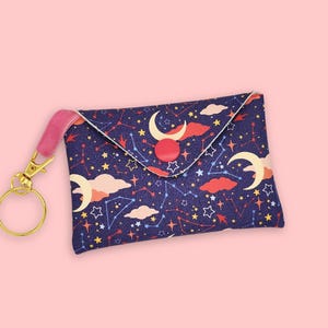 May include: A navy blue fabric wallet featuring a celestial design of moons, stars, and constellations. It has a pink strap, gold-coloured hardware, and a snap closure. The wallet is designed to hold cards and cash.