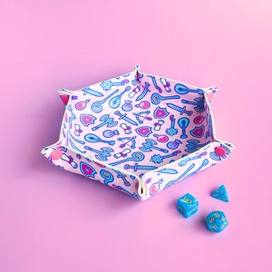 May include: A pink and white dice tray with a hexagonal shape and a pattern of blue and pink swords, shields, and other fantasy-themed items. The tray is filled with blue dice.
