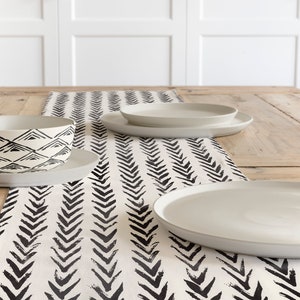 May include: A dining table setting featuring a cream-colored bowl and plates. The bowl has a black geometric pattern. A black and white arrow pattern table runner is also present, adding a decorative touch to the table setting.