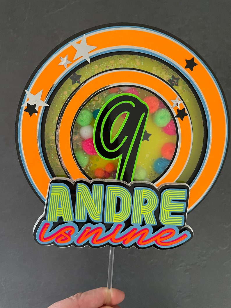 May include: A cake topper with a neon green number nine inside a circle with orange, blue, and yellow rings. The text "ANDRE is nine" is below the circle in neon green and blue letters.