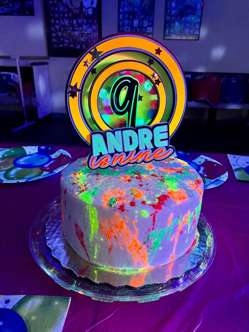 May include: A white cake with neon green, orange, and red paint splatters. The cake is topped with a yellow and black cake topper that says "ANDRE is nine" and has a large number nine in the center.