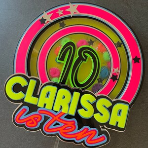 May include: A neon green and pink cake topper with the number 10 in the center, surrounded by stars and colorful pom-poms. The text "Clarissa is ten" is written in a bold, neon green font.