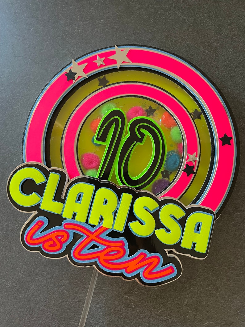 May include: A neon green and pink cake topper with the number 10 in the center, surrounded by stars and colorful pom-poms. The text "Clarissa is ten" is written in a bold, neon green font.