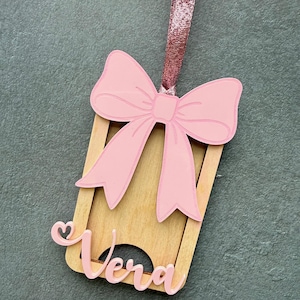 May include: A wooden tag with a pink bow and the name "Vera" in cursive. The tag has a rectangular shape with a cutout at the bottom. A pink glitter ribbon is attached to the top for hanging. The bow is a light pink color.