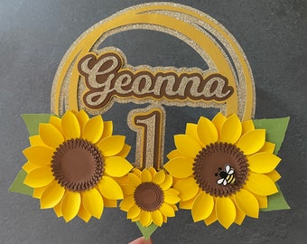 May include: A gold glitter cake topper with the name "Geonna" and the number "1" in the center. The topper is decorated with three paper sunflowers, one with a small bee.