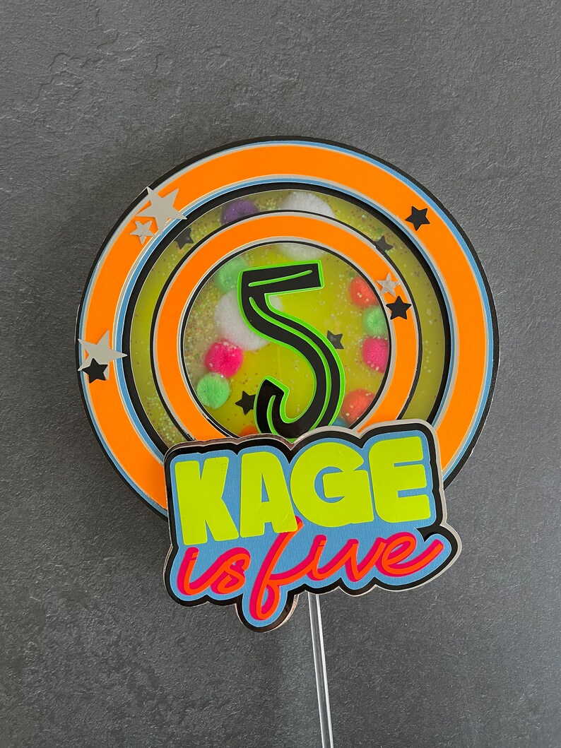 May include: A cake topper with a colorful, layered circle design featuring a green number "5" and the text "Kage is five". The topper is on a clear plastic stick.