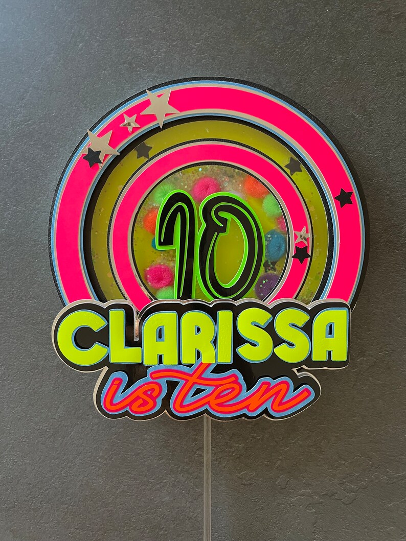May include: A colorful cake topper with a neon green number 10 in the center, surrounded by a pink and yellow circle with black stars. The text "Clarissa is ten" is written in neon green and black letters below the circle.