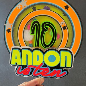 May include: A colorful cake topper with a neon green and yellow "ANDON is ten" design. The topper features a circular design with a black number "10" in the center, surrounded by a multi-colored ring.