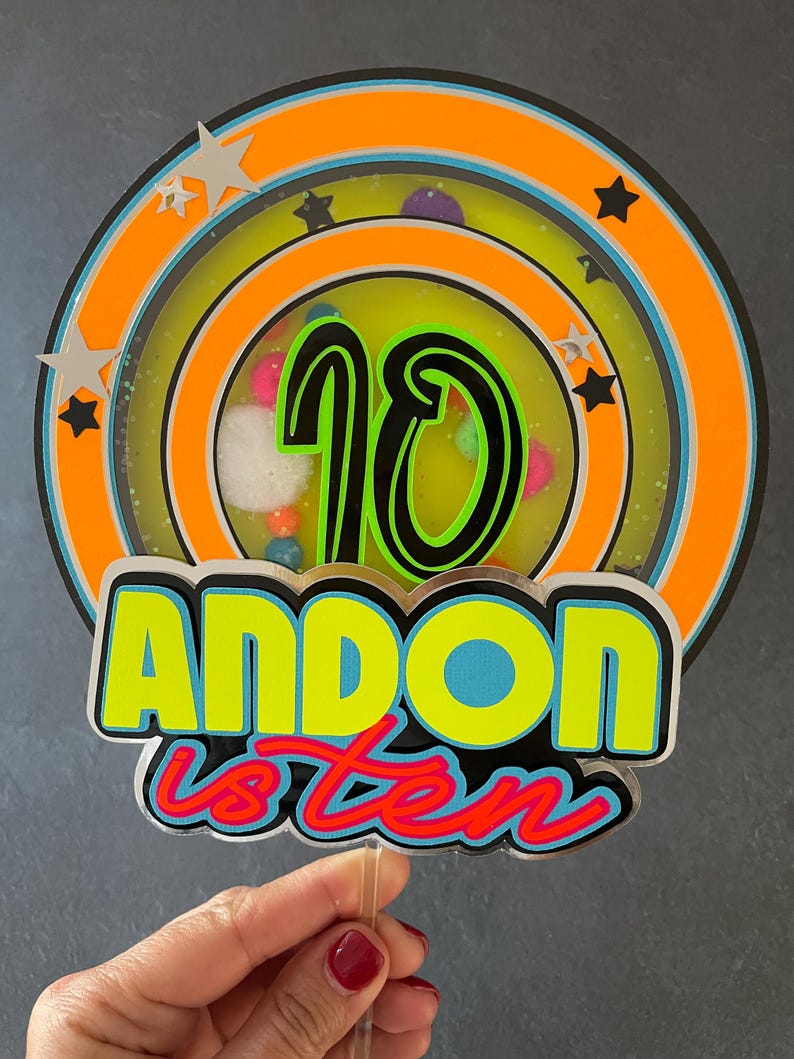 May include: A colorful cake topper with a neon green and yellow "ANDON is ten" design. The topper features a circular design with a black number "10" in the center, surrounded by a multi-colored ring.