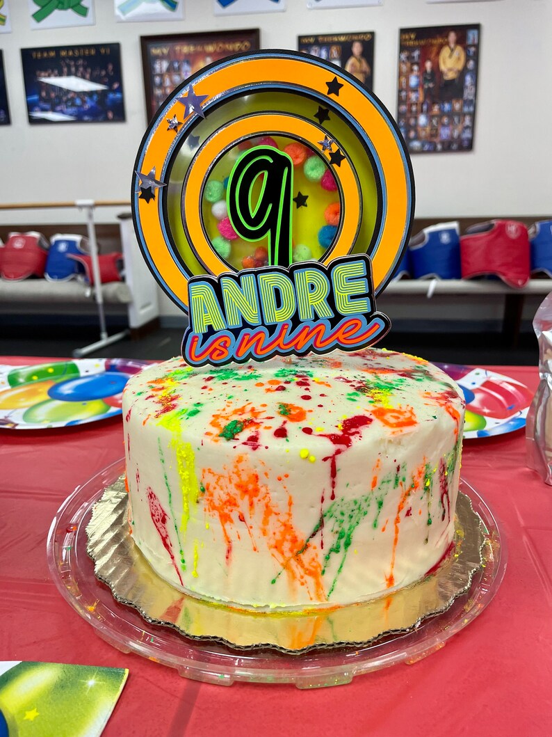 May include: A white cake decorated with colorful paint splatters. The cake is topped with a yellow and green cake topper that says "Andre is nine" and features a number nine in a circle.