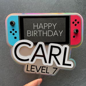 May include: A cake topper featuring a blue and red Nintendo Switch design with the text "Happy Birthday Carl Level 7".
