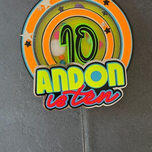 May include: A colorful cake topper with a large number 10 in the center, surrounded by a circle of stars and a ring of colorful dots. The text "ANDON is ten" is written below the number 10.