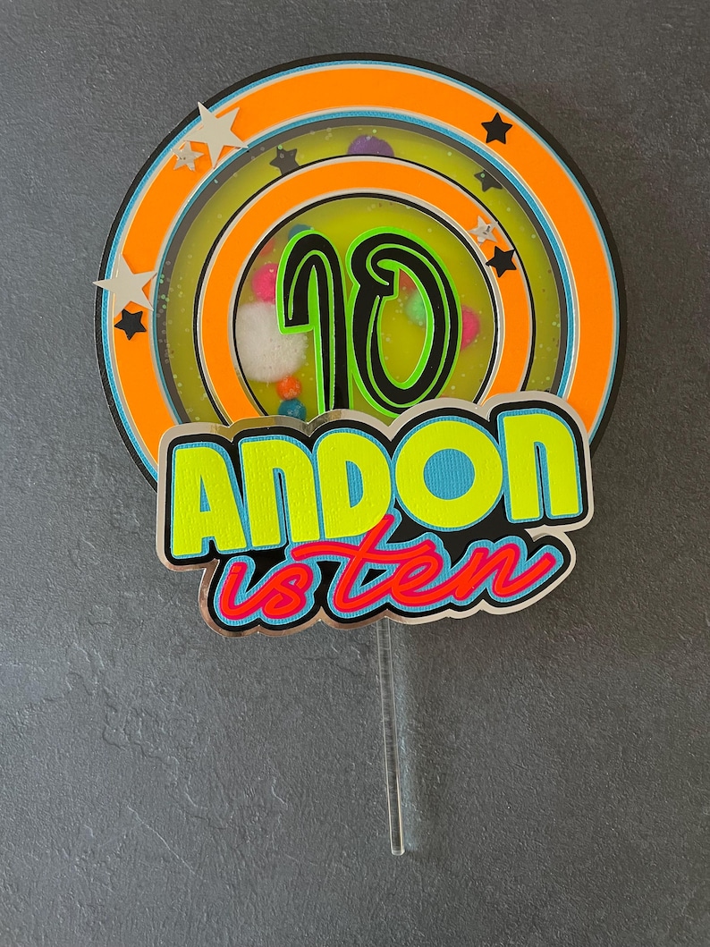 May include: A colorful cake topper with a large number 10 in the center, surrounded by a circle of stars and a ring of colorful dots. The text "ANDON is ten" is written below the number 10.